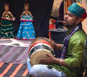 Photo of an Indian street performer hitting a drum and mid-singing surrounded in tapestries and Indian dolls