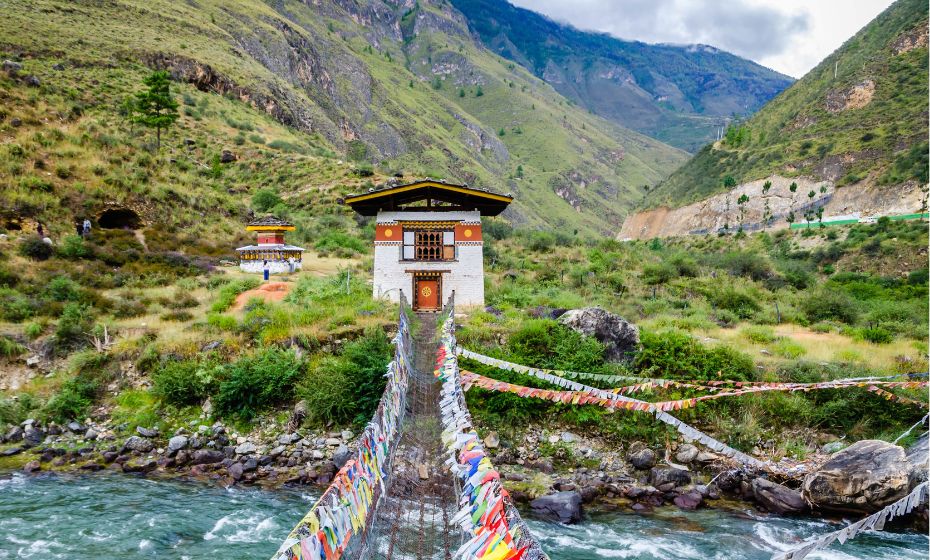 Customs and traditions in Bhutan - Iron Chain Bridge of Tamchog Lhakhang Monaster over the Paro River in Bhutan. Colourful flags adorn the sides of the wooden swinging bridge and towards a small hut-like building, and mountains sit in the backdrop of the photo.